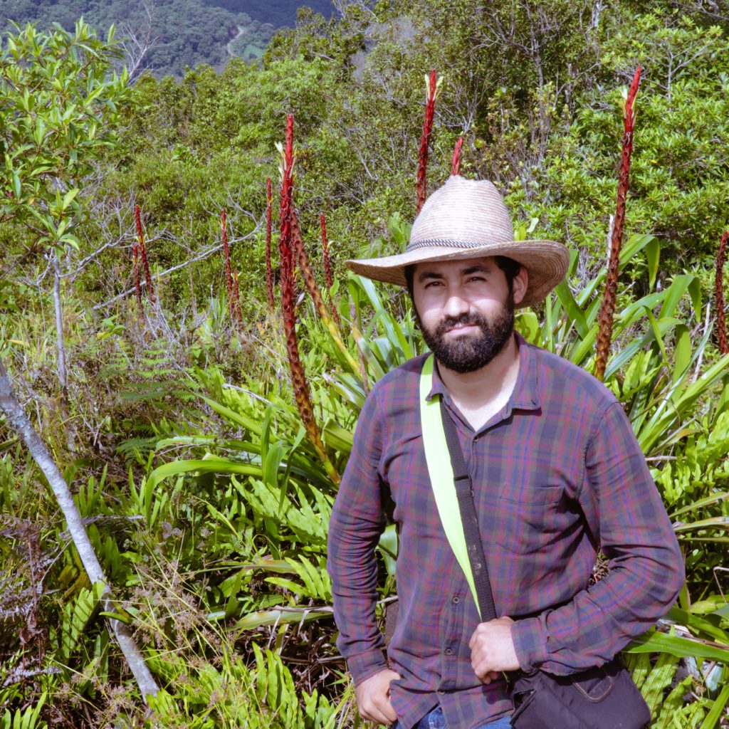 A photo of the speaker shows a thick-bearded man in a straw hat, long-sleeve purple and red plaid shirt, and blue jeans standing on a mountainside. He stares at the camera with a relaxed posture, the hat shading his eyes from the full sun on his face. He has a bag slung over his shoulder and across his chest, with his left hand grabbing the strap in front of his left hip and his right thumb hooked into his front right pants pocket. The plants behind him include ferns and light-green rosette plants with tall red flowering stalks. The background is filled with lush vegetation.