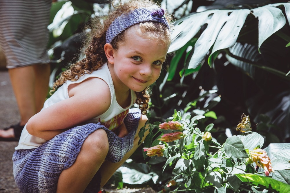 A child grins at the camera next to a butterfly balanced on a leaf.