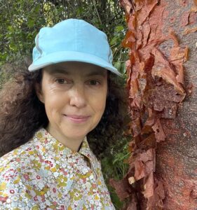Dr. Ina Vandebroek stands near a tree trunk with peeling reddish papery bark. She is wearing a turquoise hat and a flower print collared shirt. Her hair is long, brown, and curly.