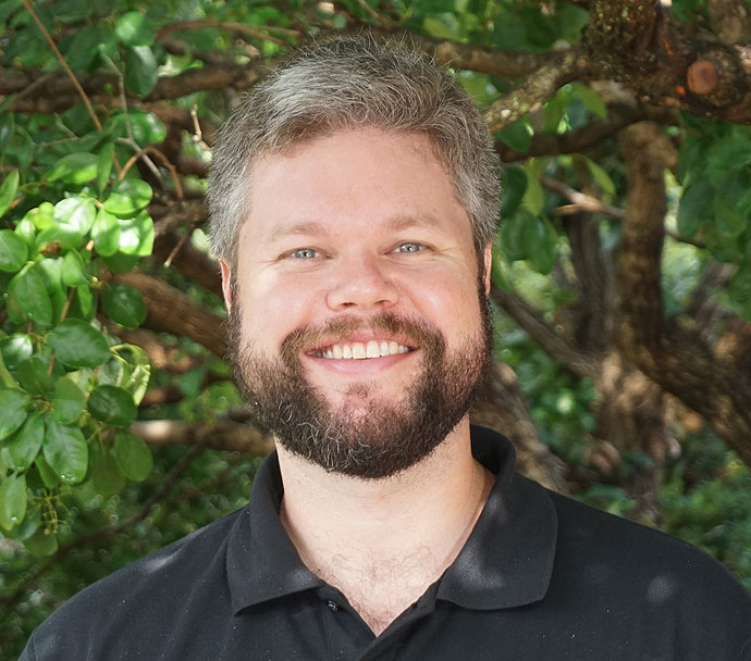 A white man with salt and pepper colored hair and a dark beard and wearing a dark, collared shirt stands smiling in front of green foliage