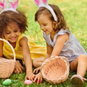 Two young girls wearing bunny ears smile as they collect colorful Easter eggs on a sunny lawn, each with a wicker basket beside them.
