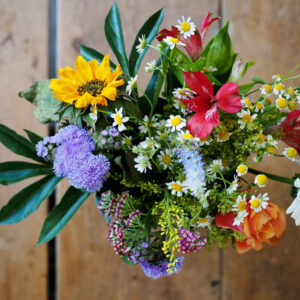 A colorful mixed bouquet featuring sunflowers, chamomile, red alstroemeria, purple ageratum, an orange rose, and greenery, viewed from above against a rustic wooden surface.