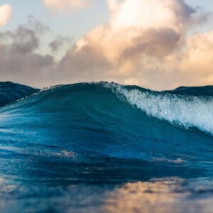 A deep blue ocean wave crests and begins to break against a sky of warm golden clouds at sunset.
