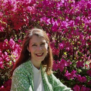 Author Mary Frances Baker smiles in a green floral jacket in front of vibrant pink azalea blooms on a sunny day.