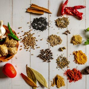 Flat lay of whole and ground spices on a white wooden surface, including ginger, garlic, green and red chilies, turmeric, cumin, coriander, black pepper, cinnamon, bay leaves, cardamom, and fresh cilantro.